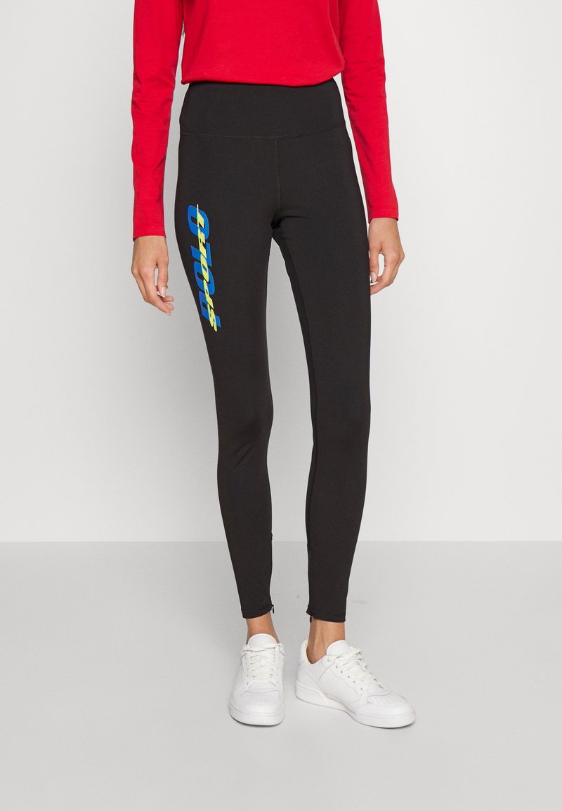 Svarta leggings i stretchtyg med hög midja och en blå- och gulllogotypgrafik på vänster ben. Vita sneakers.