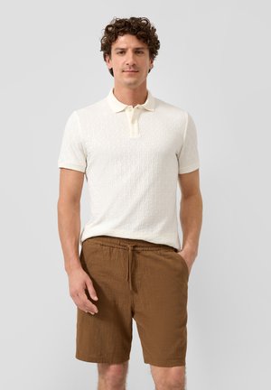 Mann steht mit Händen in den Taschen, trägt ein strukturiertes weißes Poloshirt und braune Shorts mit Kordelzug vor schlichtem grauem Hintergrund.
