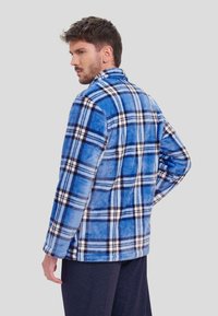 Chaqueta de forro polar a cuadros azules con una textura suave, con rayas blancas y marinas. Diseño con cuello, mangas largas y un corte holgado.