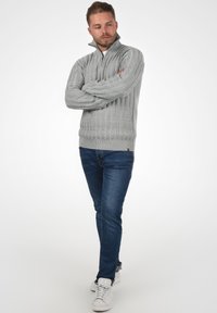 Pull à col zippé gris clair avec une texture en maille torsadée, poignets et ourlet côtelés, assorti à un jean bleu et des baskets blanches.