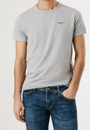 Mand iført lysegrå kortærmet t-shirt med lille sort "Pepe Jeans London" logo og blå denimjeans, den ene hånd i lommen, på hvid baggrund.