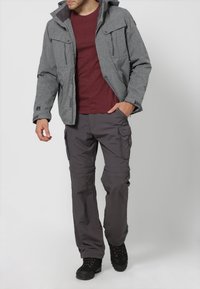 Grau strukturierte Jacke mit Kapuze, zwei Fronttaschen; weinrotes Hemd darunter; graue Cargohosen mit mehreren Taschen; schwarze Stiefel.