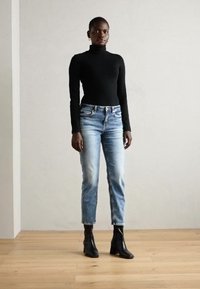 Schwarzer Rollkragenpullover, hellblaue Jeans und schwarze Ankle Boots. Das Model steht auf einem Holzboden vor einem hellen Wandhintergrund.