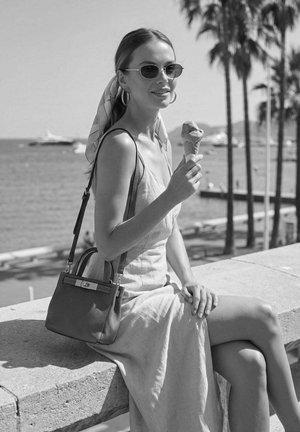 Femme en lunettes de soleil et robe assise au bord de la mer, avec des palmiers, tenant un cornet de glace et portant un sac à bandoulière.