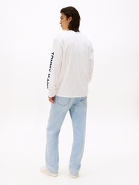 Biała koszula z długim rękawem z napisem "TOMMY JEANS" w granatowym kolorze na lewym rękawie, w połączeniu z jasnoniebieskimi dżinsami i białymi trampkami.