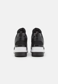 Černé tenisky s hladkým svrškem kombinujícím síťovinu a lakované detaily, bílou stélkou a robustní černou podrážkou. Dvojité poutka na patě pro snadné obouvání.