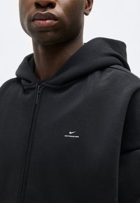 Κοντινό πλάνο ενός ατόμου που φοράει μαύρο φούτερ με φερμουάρ Nike, με μικρό λευκό λογότυπο και κείμενο "NIKE STANDARD ISSUE" στο στήθος.