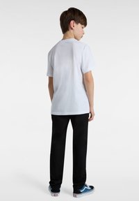 Weißes Baumwoll-T-Shirt mit kurzen Ärmeln, kombiniert mit schwarzen Hosen und blauen Sneakers, von hinten betrachtet.