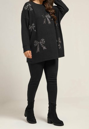 Kvinde iført sort oversized sweater med sølv paliet sløjfe mønstre, sorte leggings og sorte ankelstøvler med hæl står mod en neutral baggrund.