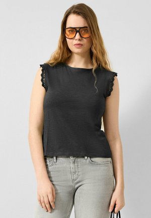 Jeune femme portant de grandes lunettes de soleil ambrées, un haut noir sans manches avec une bordure en dentelle et un jean gris clair, tenant un petit sac noir.