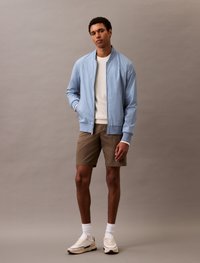 Giacca bomber azzurro chiaro con chiusura a zip, polsini a coste e un maglione bianco sotto. Pantaloncini marroni e sneakers crema completano l'outfit.