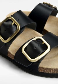 Zwarte leren sandalen met twee brede banden en gouden gespen, een kurken voetbed en een gestructureerde rubberen zool.