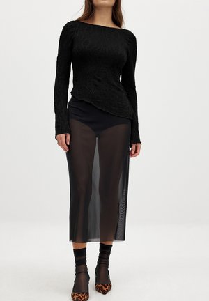 Femme portant un haut noir texturé à manches longues, une jupe midi noire transparente, des chaussettes noires et des talons pointus à imprimé léopard, posant devant un fond uni.