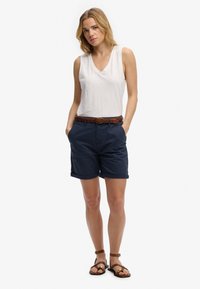 Mujer de pie con las manos en los bolsillos, vistiendo una blusa blanca sin mangas, pantalones cortos azul marino con cinturón trenzado marrón y sandalias marrones sobre fondo blanco.