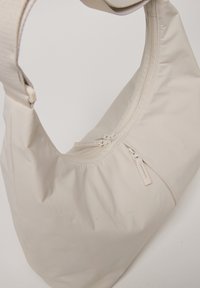 Sac à épaule beige en tissu doux avec une texture lisse. Il possède une fermeture à glissière et une large sangle unique pour le porter.