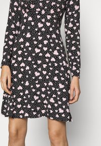 Svart klänning med långa ärmar, prydd med ett mönster av rosa hjärtan och små vita hjärtan. Tyget verkar slätt med en figurnära design och utställd kant.