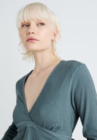 Grön texturerad topp med djup V-ringning och lång ärm, med en samlad midjedetalj. Modellen har kort, ljust hår.