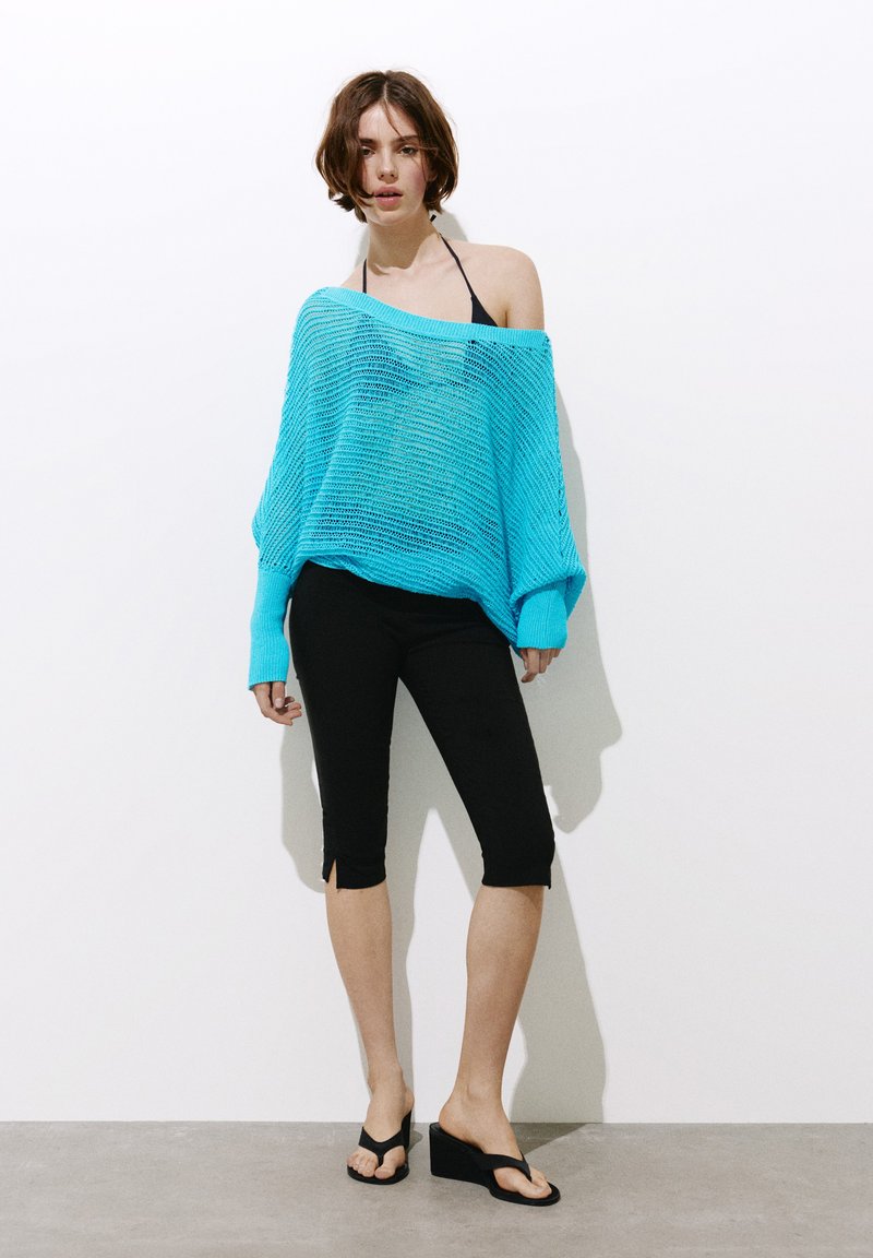 Jeune femme portant un pull en maille turquoise ample par-dessus un haut de bikini noir, des leggings courts noirs et des sandales compensées noires, debout contre un mur blanc.