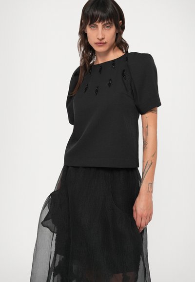 Melnas īsā piedurkņu topiņš ar kastes formu. Bezmaksas rotājumi uz priekša un mīksts, teksturēts apdare. Saskaņots ar caurspīdīgu melnu svārku.