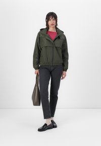 Jaqueta verde, leve, com capuz, punhos elásticos e cintura. Usada sobre uma blusa rosa, combinada com jeans escuros e mocassins pretos. Acessório com uma bolsa castanha.