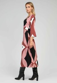 Robe maxi à motif géométrique avec des couleurs rose, noir et blanc. Présente des manches longues et une taille cintrée. Associée à des bottes noires montantes.