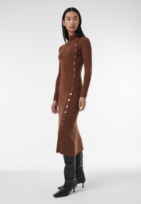 Robe en tricot côtelé marron avec un col haut, détails à boutons sur le côté et une silhouette ajustée, associée à des bottes noires jusqu'aux genoux.