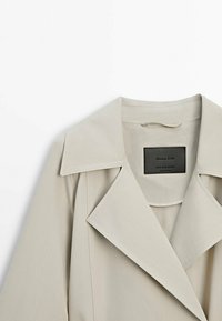 Beżowy trencz z szerokimi klapami, gładką teksturą i prostokątną metką wewnątrz. Materiał jest lekki, idealny do noszenia w warstwach.