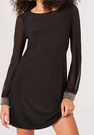 Kobieta w czarnej sukience z długim rękawem, przezroczystymi rękawami i ozdobnymi mankietami, trzymająca brzeg sukienki z jednej strony.