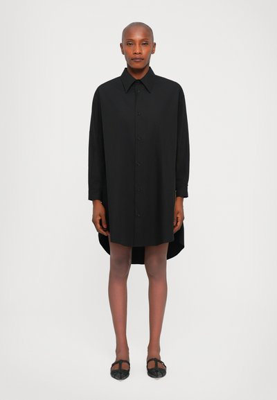 Robe chemise noire avec un col, fermeture avant par boutons, manches longues et ourlet asymétrique. Texture lisse, coupe décontractée, design mi-genoux.