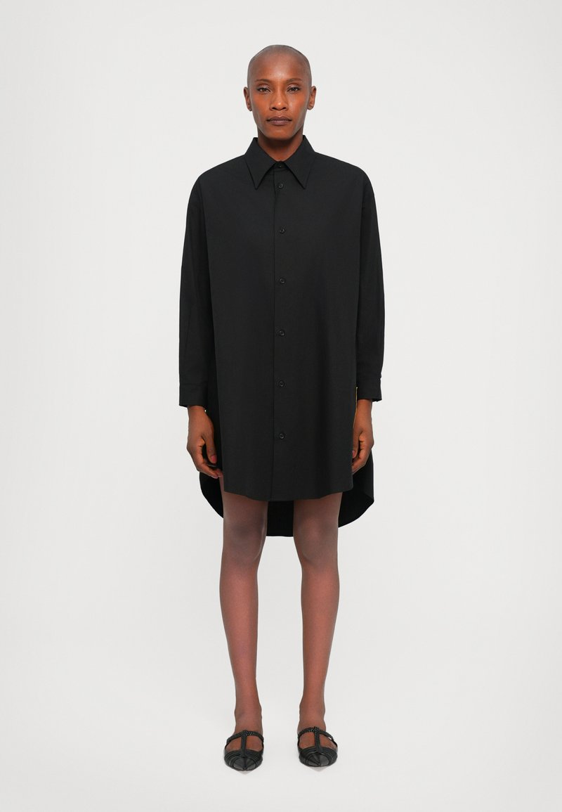 Black shirt dress with a collar, front button closure, long sleeves, and a high-low hem. Smooth texture, relaxed fit, knee-length design.