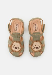 Grønne semskede sandaler med et løveansigt design på tåen, med en beige accent. Lukket bag, åbne sider og justerbare stropper.