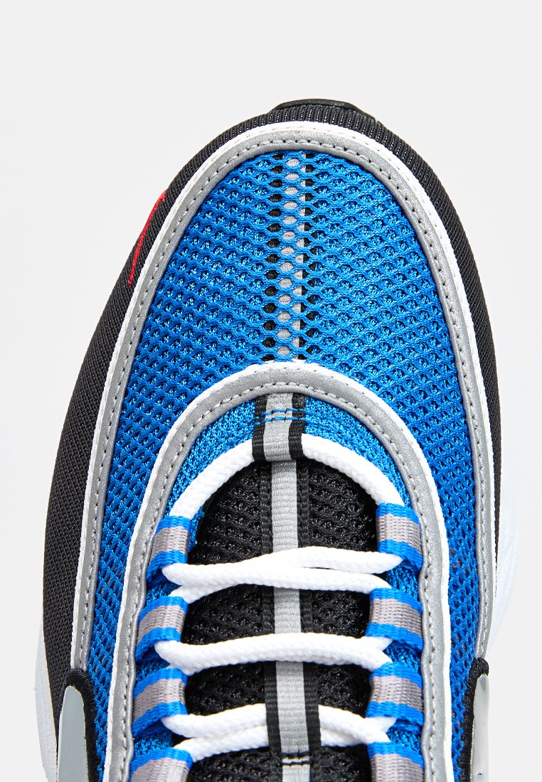 Zapato deportivo con parte superior de malla azul y detalles en negro. Cuenta con cordones blancos y grises, punta redondeada y superficie texturizada.