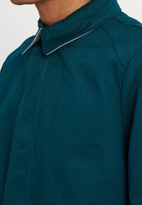 Hombre vestido con una chaqueta verde azulado de cuello plano y costuras visibles, mostrado desde el cuello hasta la mitad del pecho sobre un fondo liso.