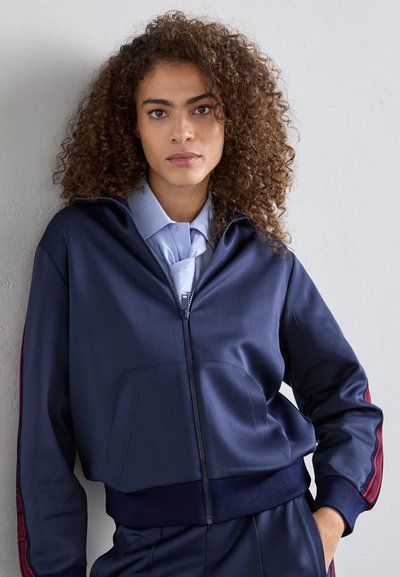 Giovane con capelli ricci che indossa una giacca a zip blu navy sopra una camicia colletto azzurro chiaro, in piedi contro un muro grigio chiaro.