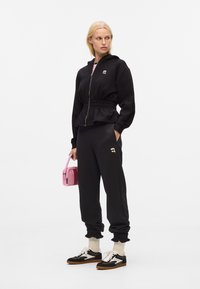 Haut à capuche noir zippé avec une taille cintrée, associé à un jogging noir. Sac à main rose notable et baskets rétro noires avec des accents blancs.