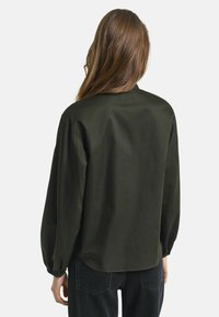 Cameretta verde scuro a maniche lunghe con vestibilità rilassata, scollo rotondo e polsini arricciati, realizzata in tessuto liscio, indossata con jeans neri.