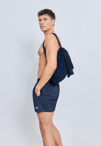 Tamsiai mėlyni šortai su raišteliais ir logotipu, derinami su atitinkančia rankine. Rankinė turi tekstūruotą paviršių ir dvi petnešėles.