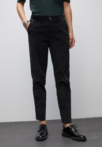 Persona che indossa pantaloni neri a sigaretta con tasche, scarpe nere stringate, top verde scuro e braccialetti d'argento sul polso sinistro.