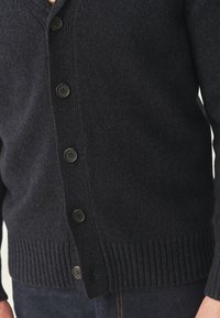 Tumši pelēks adīts kardigāns ar V veida kakla izgriezumu, sešām melnām pogām, ribotām piedurknēm un apakšmalu. Tekstūrveida audums ar gludu apdari.