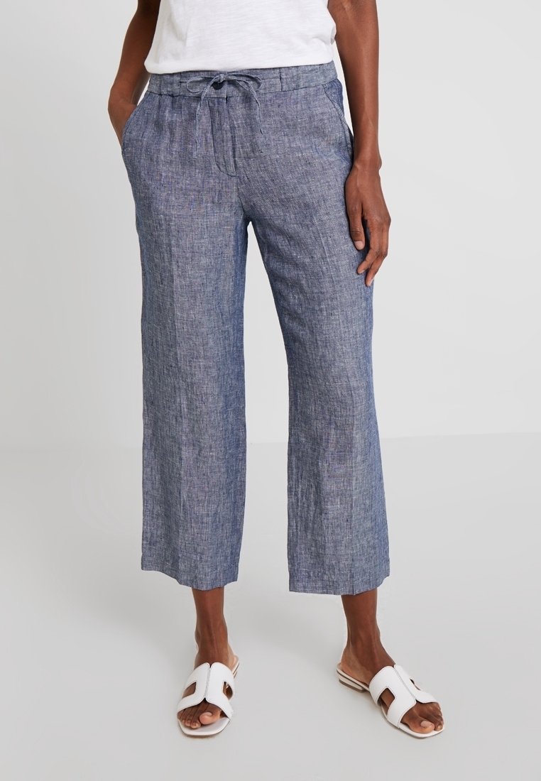 Lehké modré lněné kalhoty s uvolněným střihem, mají elastický pas na šňůrku a dvě boční kapsy. Vypadá s bílými skluznými sandály.
