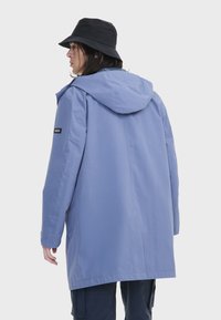 Abrigo impermeable azul con capucha, hecho de material suave. Presenta un parche de marca negro en la manga y un ajuste holgado.