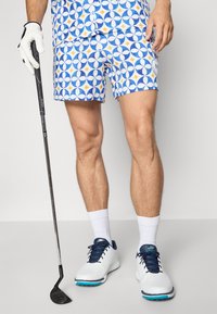 Shorts de golf avec un motif circulaire bleu et blanc agrémenté de touches jaunes. Portés avec des chaussures de golf blanches et un gant, tenant un club de golf.