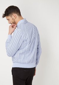 Chaqueta a rayas azul claro con puños y cuello acanalados. Presenta un diseño ajustado y un detalle de cremallera en el hombro. Simple y casual.