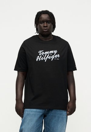 Person med mørk hud og dreadlocks iført en sort "Tommy Hilfiger" T-shirt og blå jeans mod en ensartet lys baggrund.