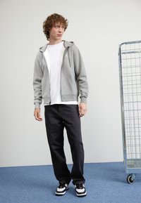 Grå zip-up hettegenser med hvit logo, paret med en hvit t-skjorte og svarte bukser. Outfiten er fullført med svart og hvit sneakers.