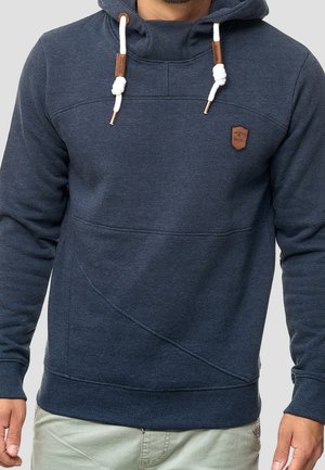 Mann trägt einen marineblauen Kapuzenpullover mit weißen Kordelzügen und einem kleinen braunen Lederpatch auf der Brust.