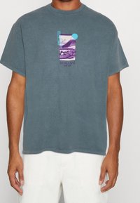 Baumwoll-T-Shirt in einem gedämpften Blau-Grau mit einem rechteckigen lila Grafik, das eine gebirgige Landschaft zeigt und text "HOKUSAI" in der Mitte.