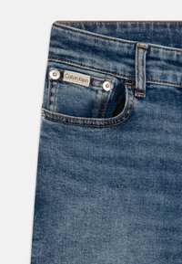 Blå denim jeans med en liten frontlomme, sølvfargede detaljer og en Calvin Klein-lapp. Stoffet har en glatt, lett teksturert overflate.