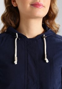 Marinblå jacka med huva, med vita snören, dragkedja framtill och textilstruktur. Inkluderar en bröstficka med sömdetalj.