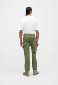 Homem com cabelo trançado vestido com uma t-shirt branca, calças cargo verde-oliva e botas beige atadas, de costas contra um fundo branco.